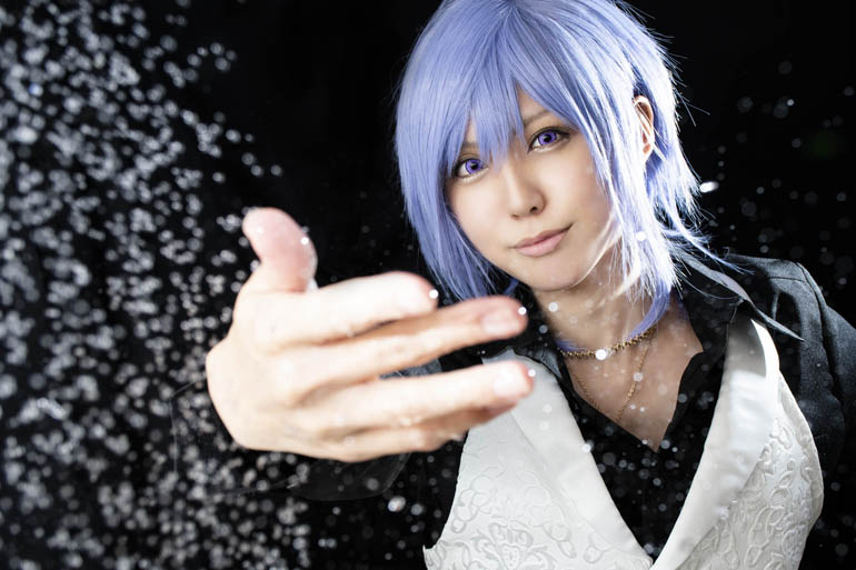 黒い布を背景に、こちらに向かって水しぶきを飛ばすように手を伸ばす男装の女性の写真。