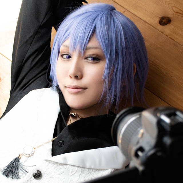 フローリングの床に寝ころび、一眼レフカメラで自撮りする男装の女性の写真。