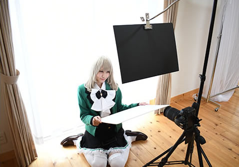 窓際で白い紙を手に持ち光を調節して写真撮影する女性の様子。