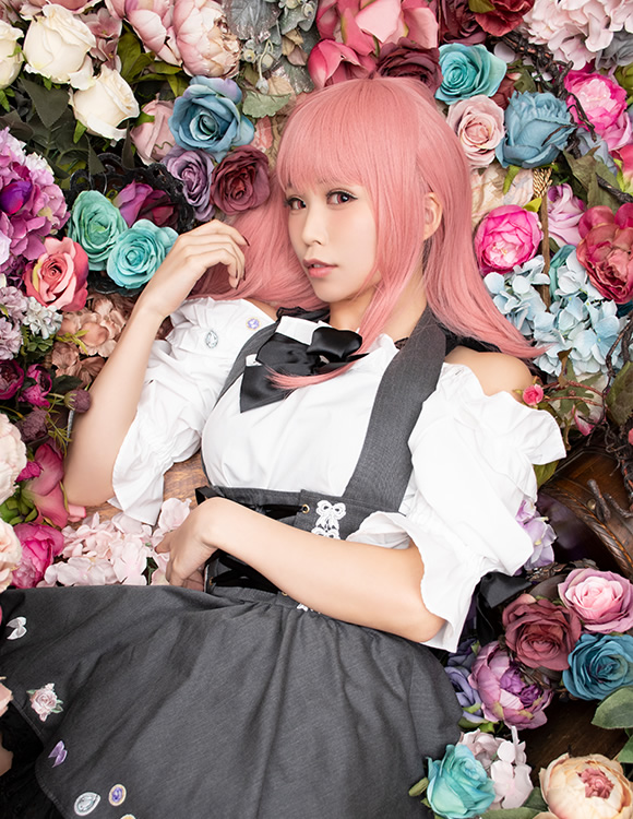 色とりどりの花の中に寝ころび、こちらを見上げる女性の写真。