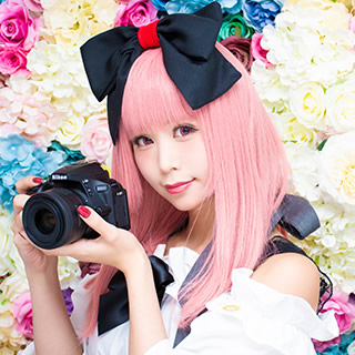 色とりどりの花を背景に、一眼レフカメラを持って微笑む女性の写真。