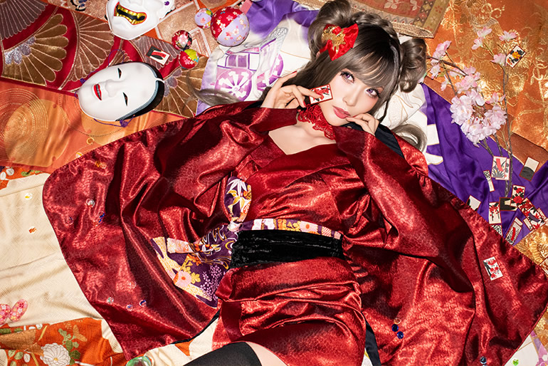 部屋中に広げた着物や帯の上に寝転び、花札を1枚手にした赤い着物の女性の写真。