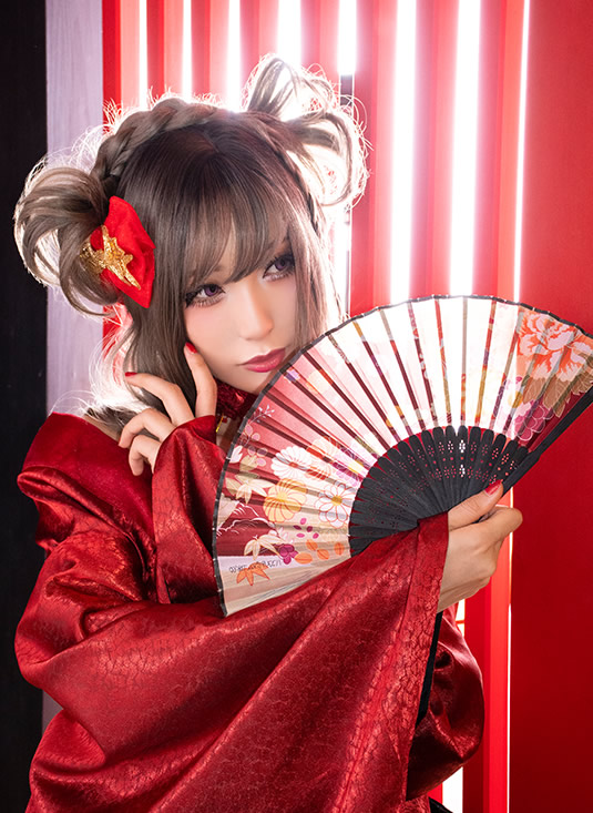 扇子を手に、赤い格子戸の傍らでポーズを取る赤い着物の女性の写真。