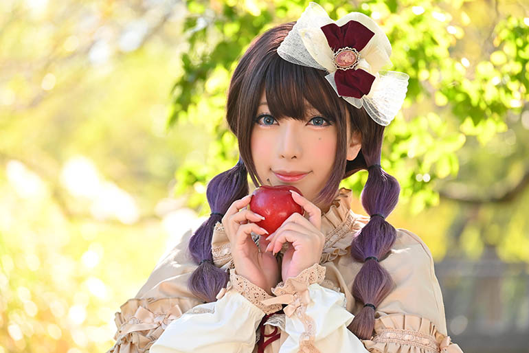 木立の下、真っ赤なリンゴを手にポーズを決める女性の写真。