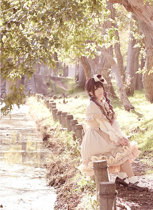 小川沿いのフェンスに腰掛けこちらを見つめる女性の写真。