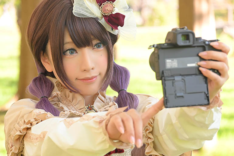 木立を背景に、カメラの画面モニターを見て「自分撮り」の操作をする女性の写真。