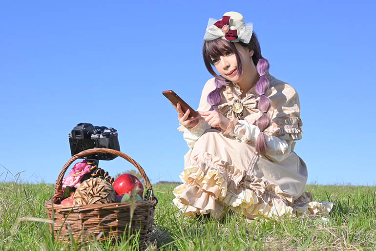 草原で三脚に立てたカメラを、手元のスマホでリモート操作して「自分撮り」する女性の写真。